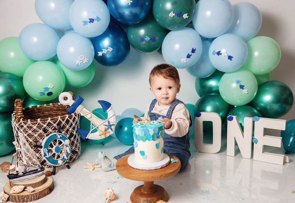First birthday celebration setup with blue and green balloon arch, cake, and 'ONE' letters against white background.