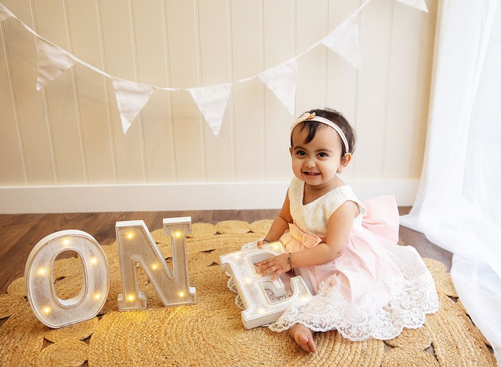 A child in a white lace dress sits on a gold carpet next to glowing 'ONE' marquee letters and white bunting decor.