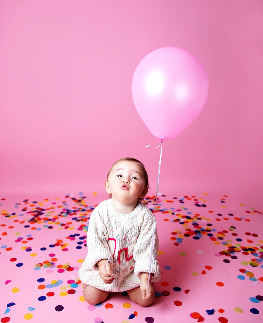 A small child in white sits on colorful confetti with a pink balloon against a rosy background.