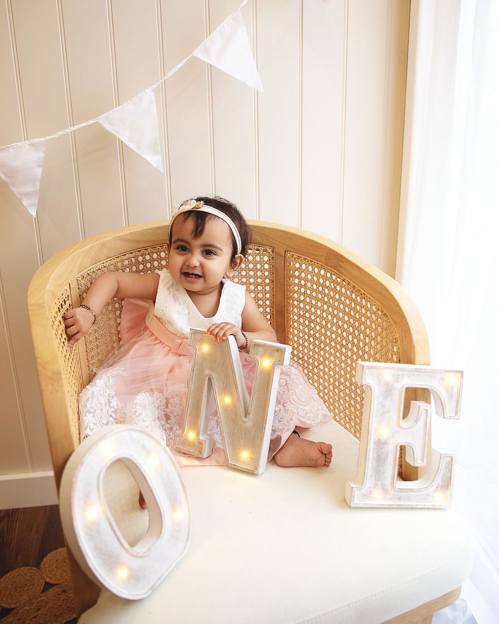 Baby in pink dress sits in vintage cane chair with decorative light-up letters and white banner bunting overhead.