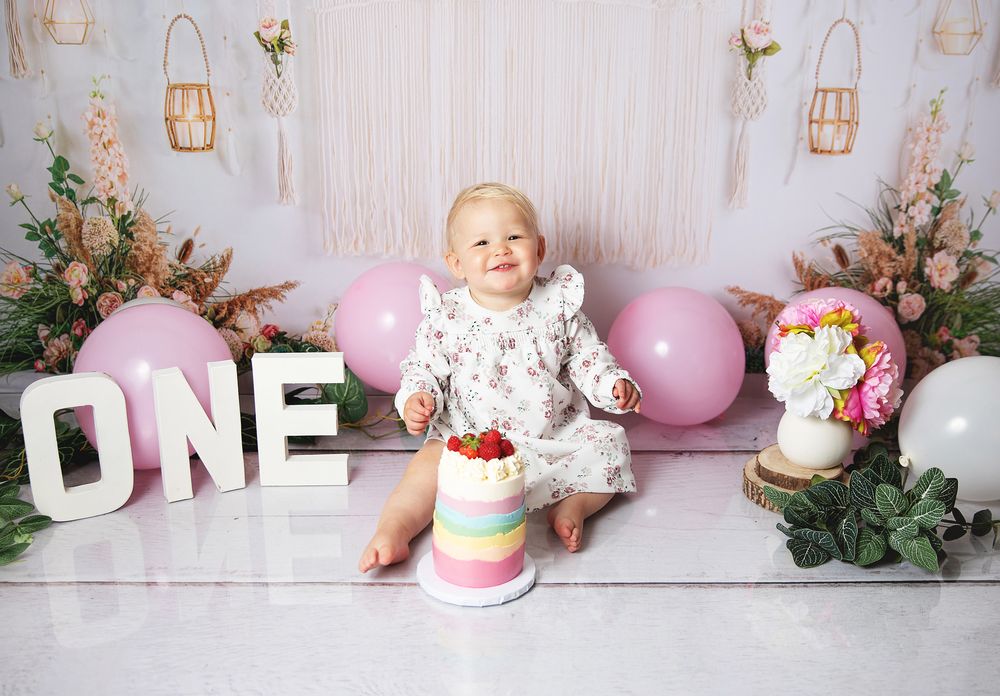 A first birthday celebration setup with 'ONE' letters, pink balloons, floral decorations and a colorful tiered cake.