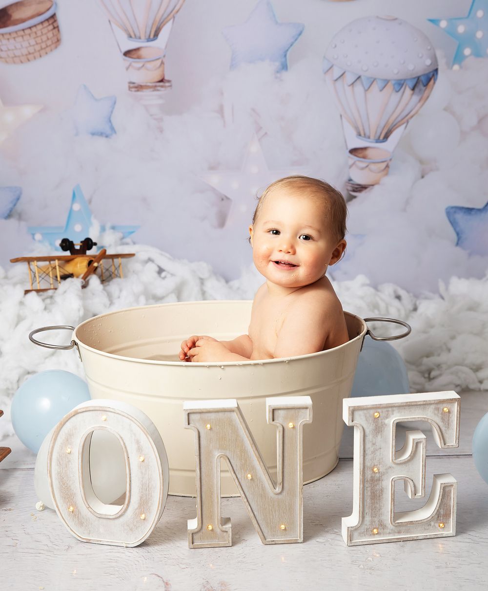 Decorated baby photography setup features a metal tub, cloud backdrop with hot air balloons, and decorative ONE letters.