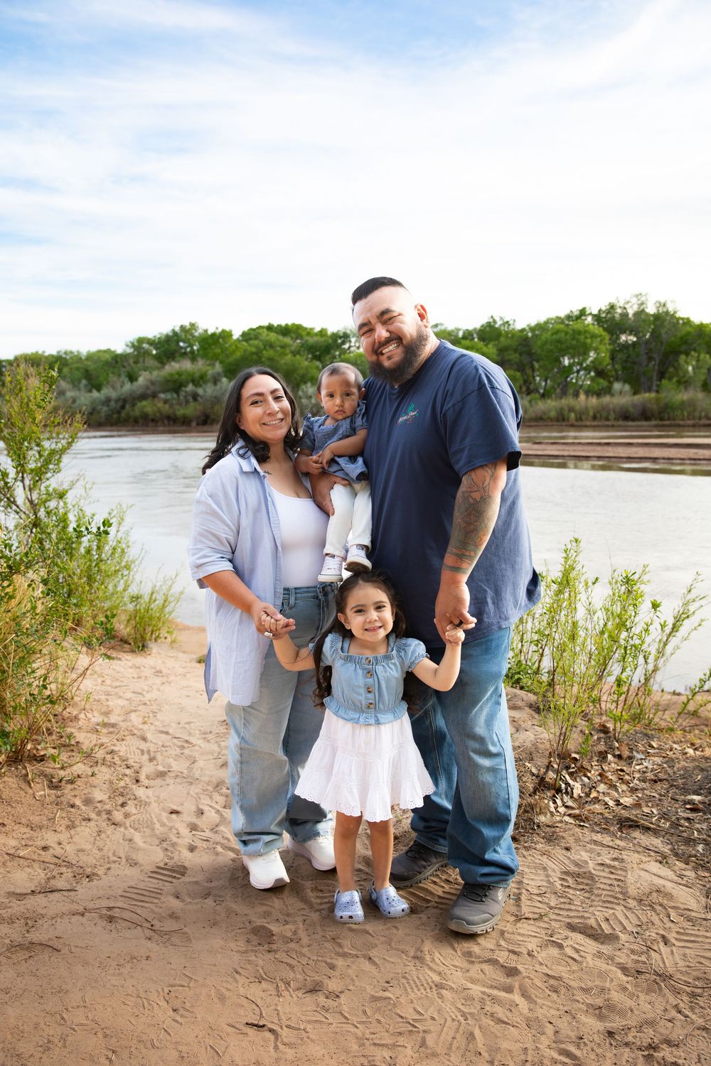 A happy family of four poses on a sandy riverbank surrounded by green trees and a cloudy blue sky.