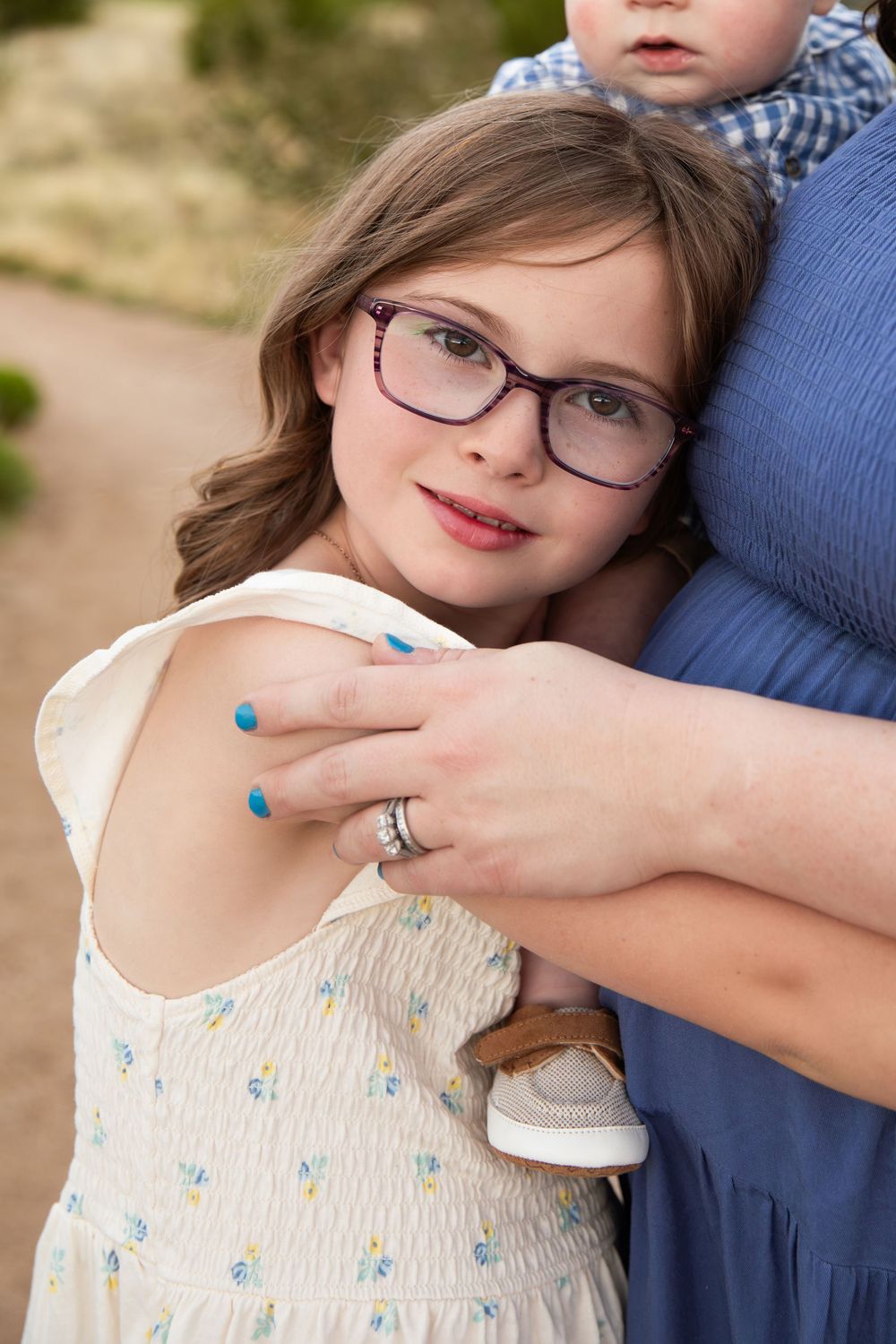 Young girl with glasses hugs her mother outdoors, while a toddler rests on mom's hip in a heartwarming family moment.