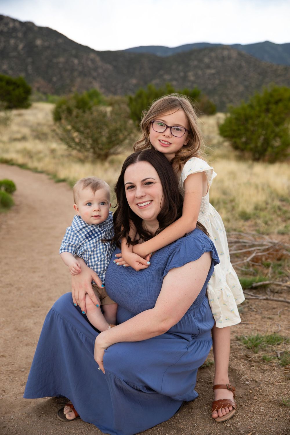 A mother in a blue dress kneels outdoors with her toddler son and young daughter against a mountain backdrop.