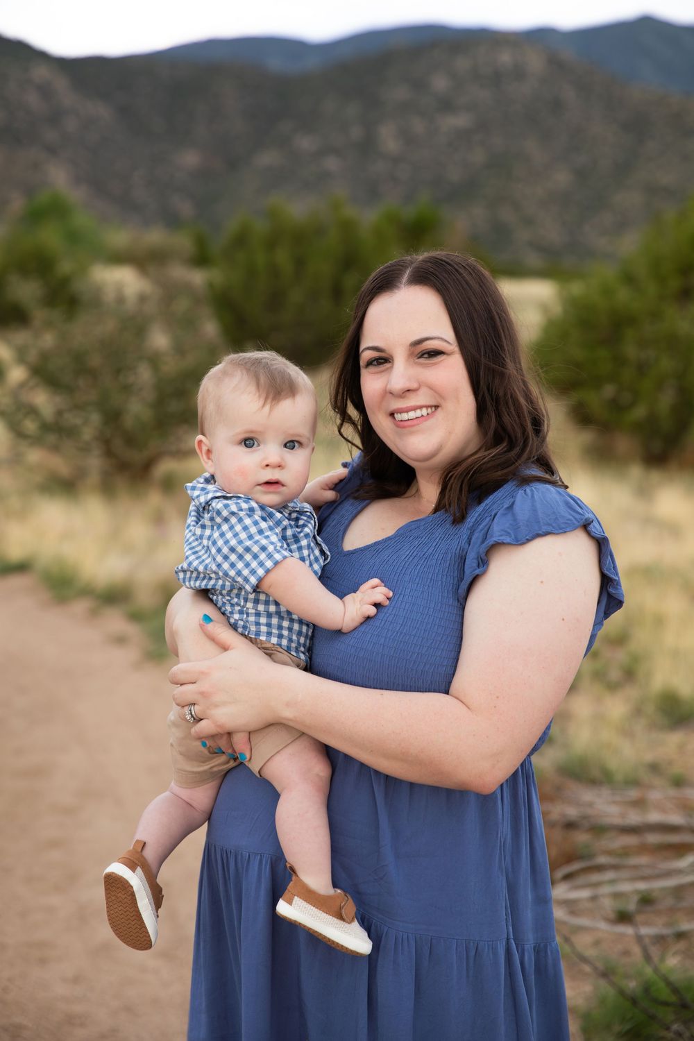 A smiling woman in a blue dress holds a baby in a checkered outfit outdoors with mountains in the background.