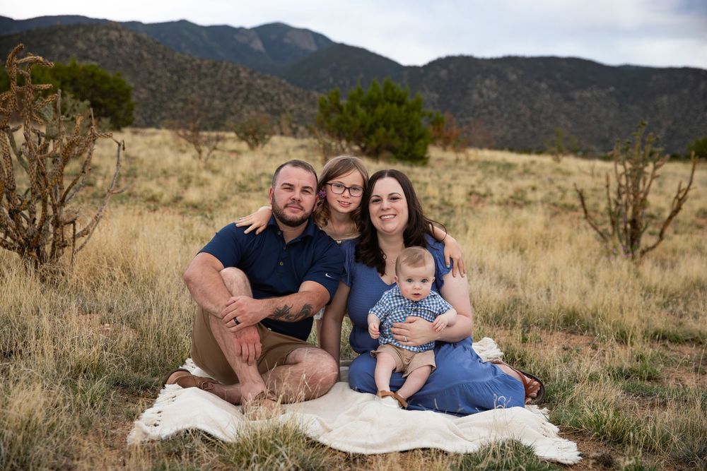 Family of three sitting on blanket in mountain meadow, parents smiling with baby in blue outfit outdoors.