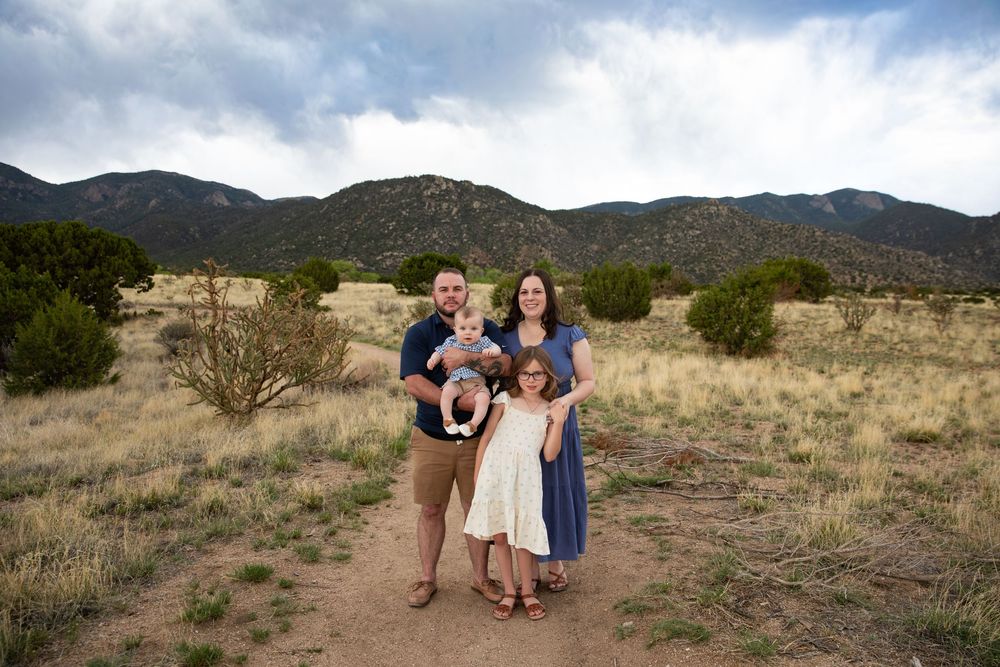 Family of four poses on a desert trail with mountain backdrop under cloudy skies in New Mexico.
