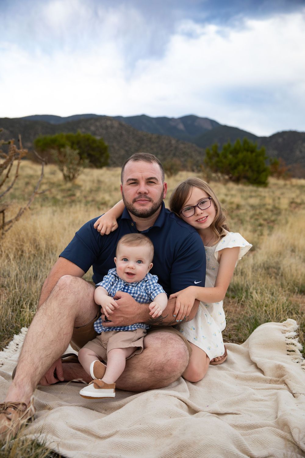 A father sits on a blanket outdoors holding a baby, with a young girl in glasses beside him, mountains in background.