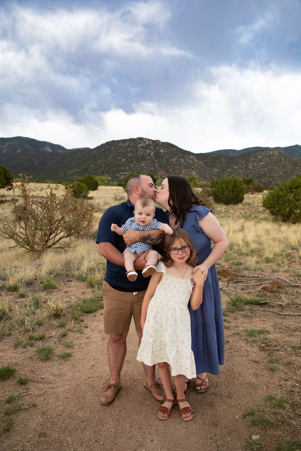 Family of four poses outdoors with mountain backdrop, parents kissing baby while young girl smiles in floral dress.