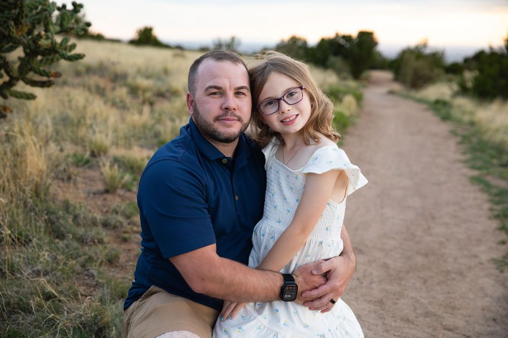 A man and young girl with glasses share a warm embrace on a scenic dirt path surrounded by golden meadows.