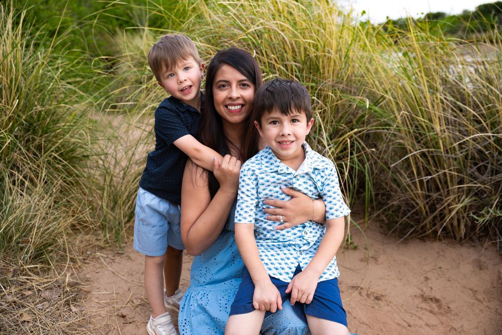 A woman smiles with two young boys on a sandy beach surrounded by tall green grass.