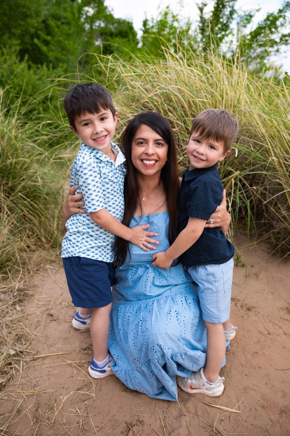 A smiling mother in a blue dress kneels with her two boys hugging her on a sandy path.
