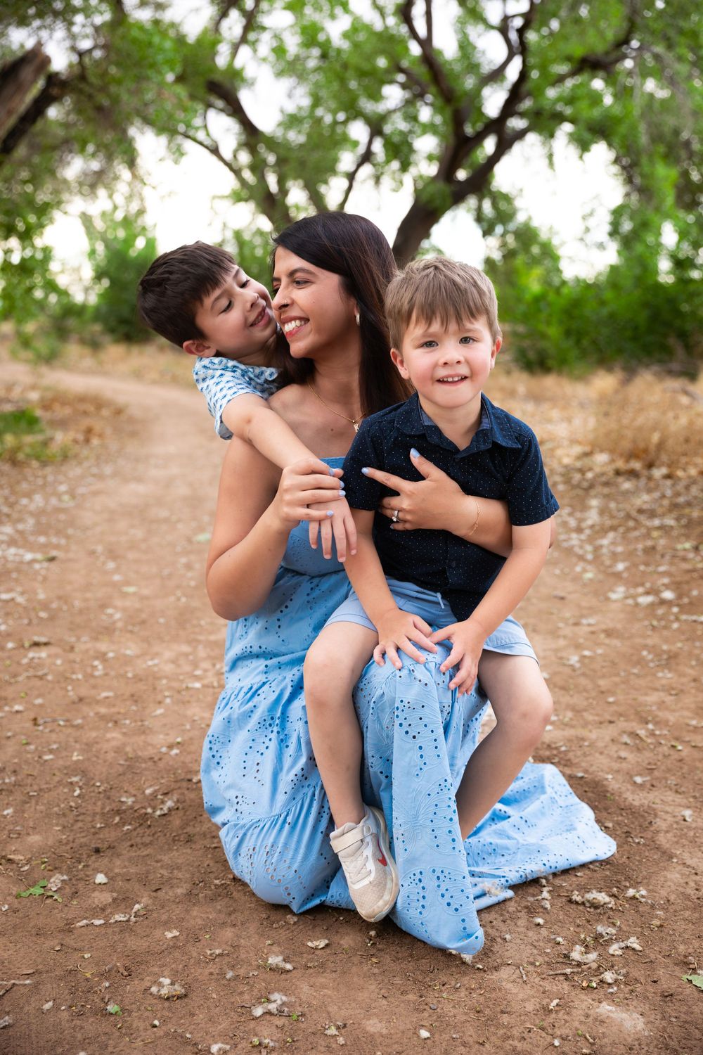 A mother in a blue dress kneels on a dirt path hugging her two young sons outdoors.