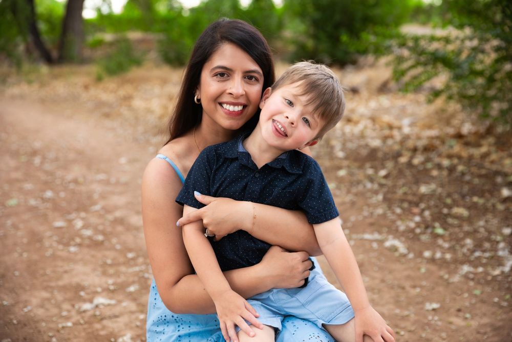 A smiling woman hugs a laughing young boy on a dirt path surrounded by green trees outdoors.