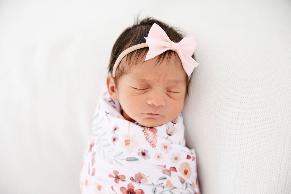 Sleeping newborn baby swaddled in a floral blanket wearing a pink bow headband on a white background.