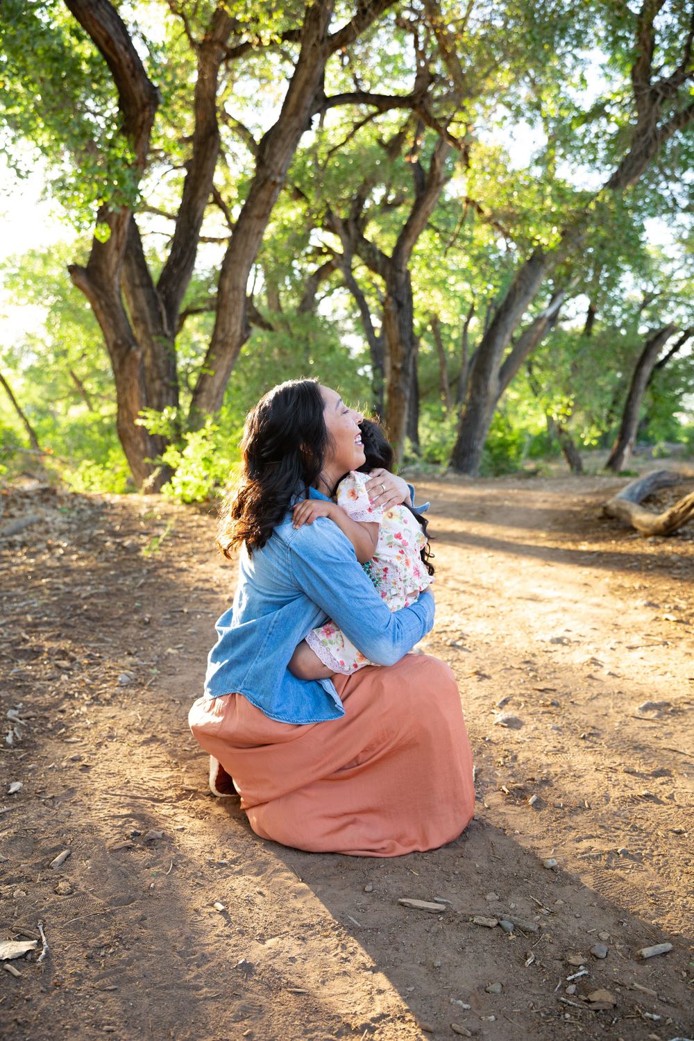 A mother kneels on a dirt path hugging her child in a sunlit forest with lush green trees.