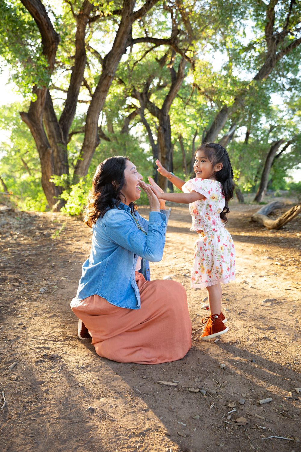 A mother kneels on a dirt path playing with her young daughter in a sunlit forest park.