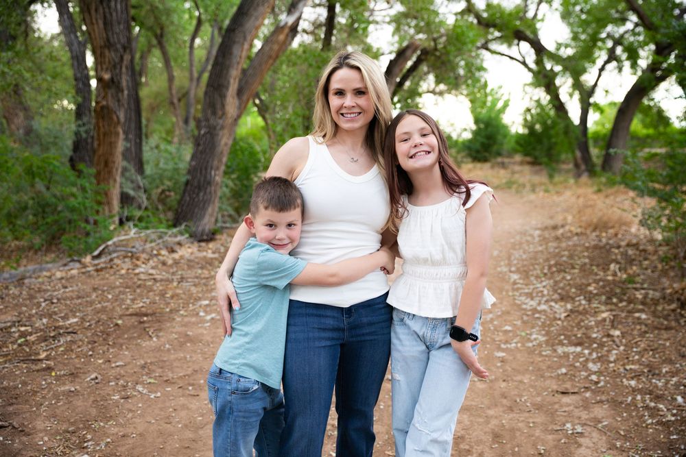 A woman and two children smile together on a wooded dirt path, posing for a family portrait outdoors.