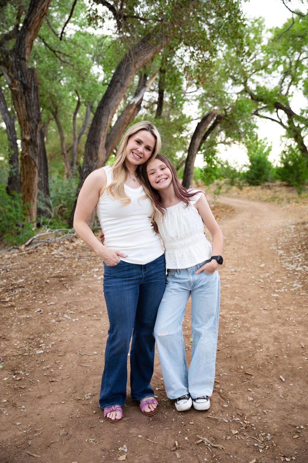 A smiling woman and young girl in white tops and jeans pose together on a dirt path surrounded by trees.