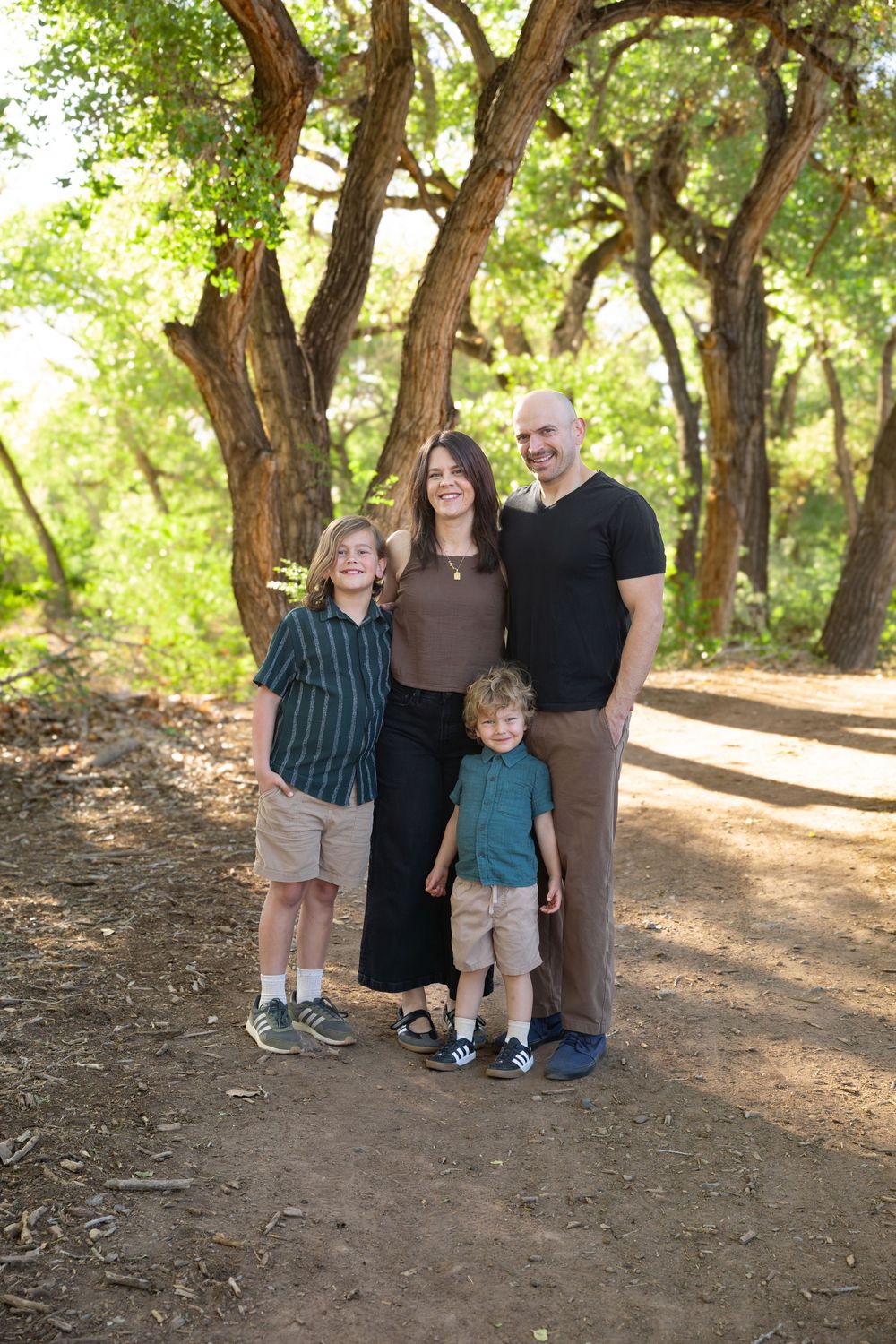 Family of four posing on a shaded dirt path surrounded by lush green trees in a park setting.