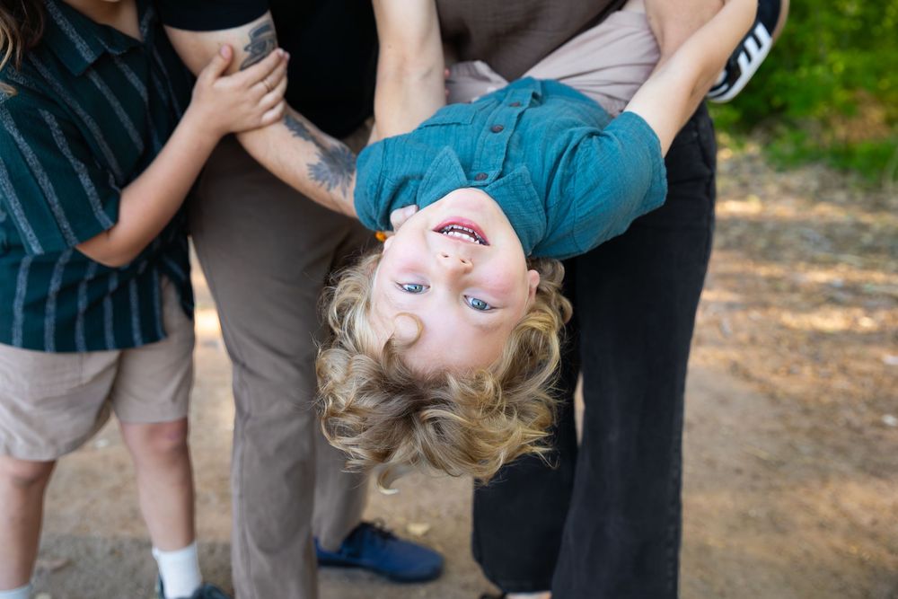 A laughing blond child being held upside down by adults outdoors, showing joy and playfulness during family fun.