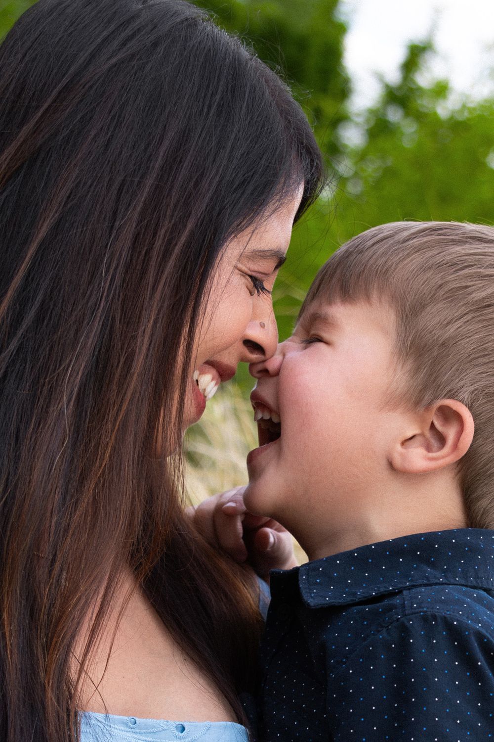 A young mother and her laughing toddler son share a playful nose-to-nose moment outdoors.