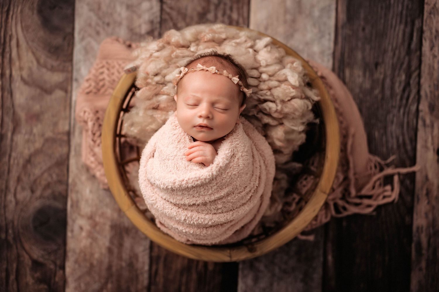 A sleeping newborn swaddled in a soft pink wrap rests peacefully in a round wooden bowl against a dark backdrop.