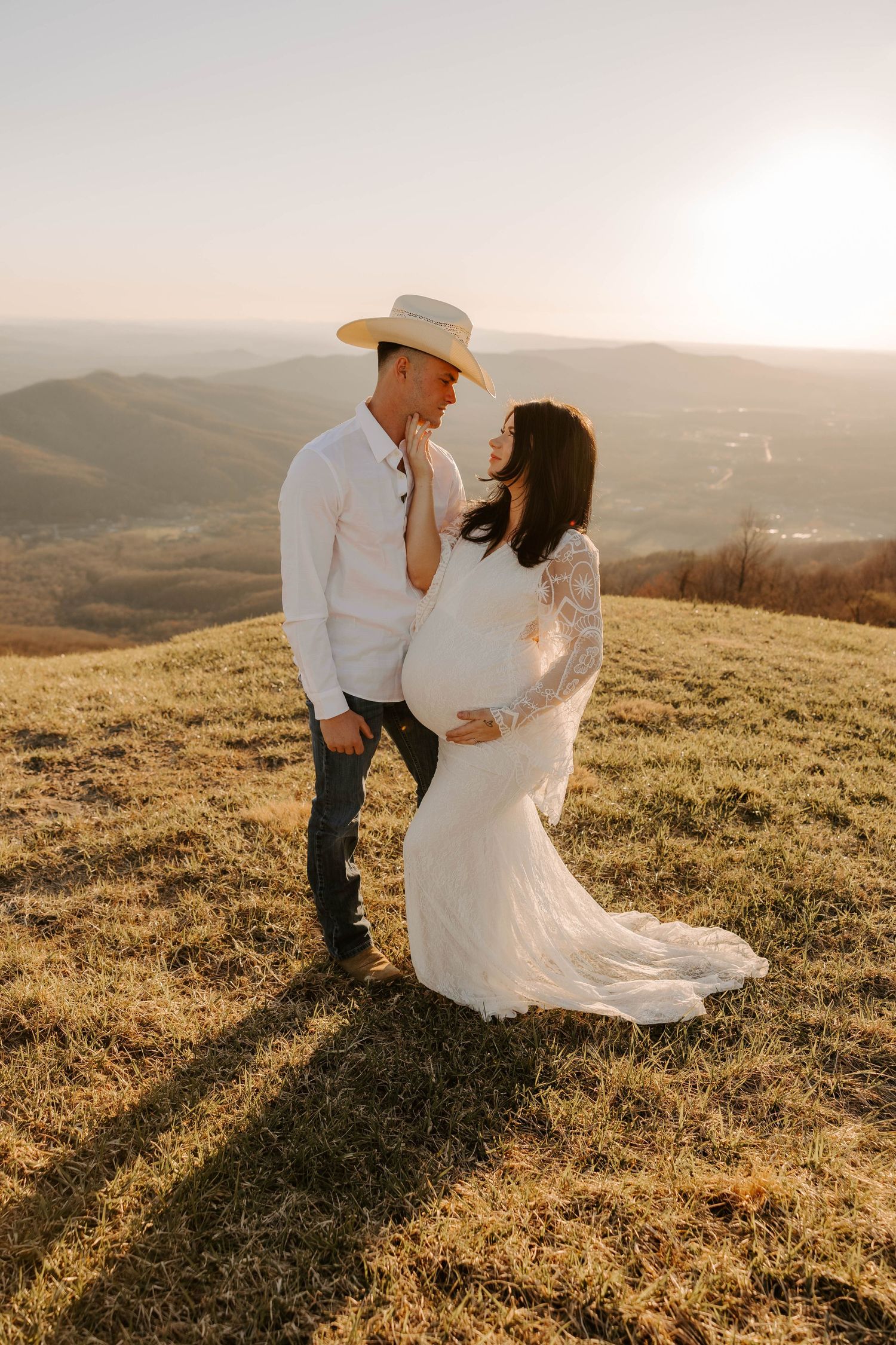 Woman in flowing white lace dress twirls in sunlit meadow during maternity shoot.