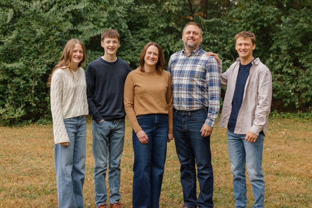 Family of five smiling outdoors on grass with lush green trees in background, wearing casual fall attire.