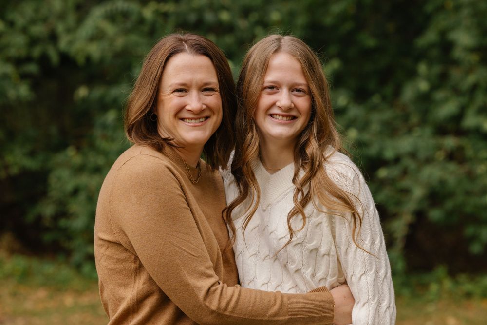 A smiling woman and young girl pose together outdoors, wearing warm tones against a lush green background.