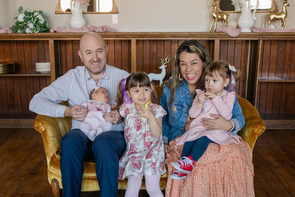 Happy family of five posing together on a gold velvet chair, parents holding three young daughters in a warmly decorated room.