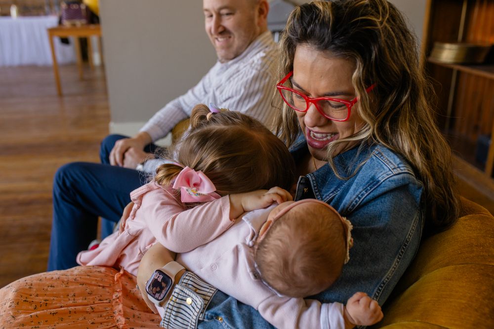 A woman with red glasses holds a newborn baby while a toddler hugs her, as a man smiles in the background.