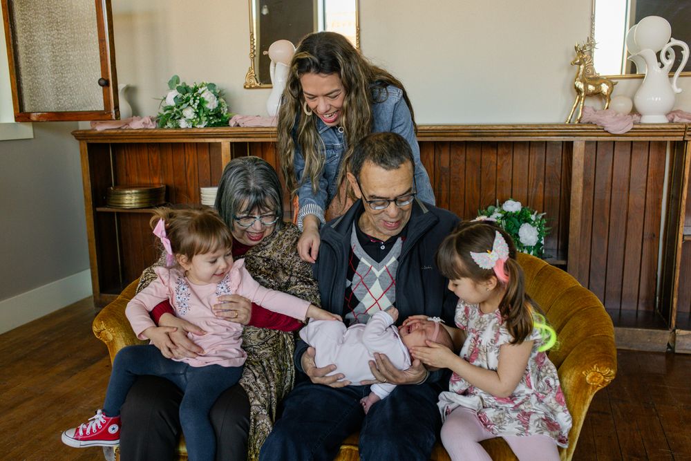 Family gathered around newborn baby, with grandparents and young girls smiling together in a warmly decorated living room.
