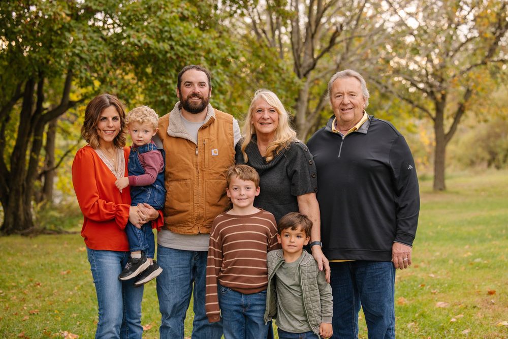 Family of seven posing outdoors in fall foliage, wearing casual autumn attire in a lush green park setting.