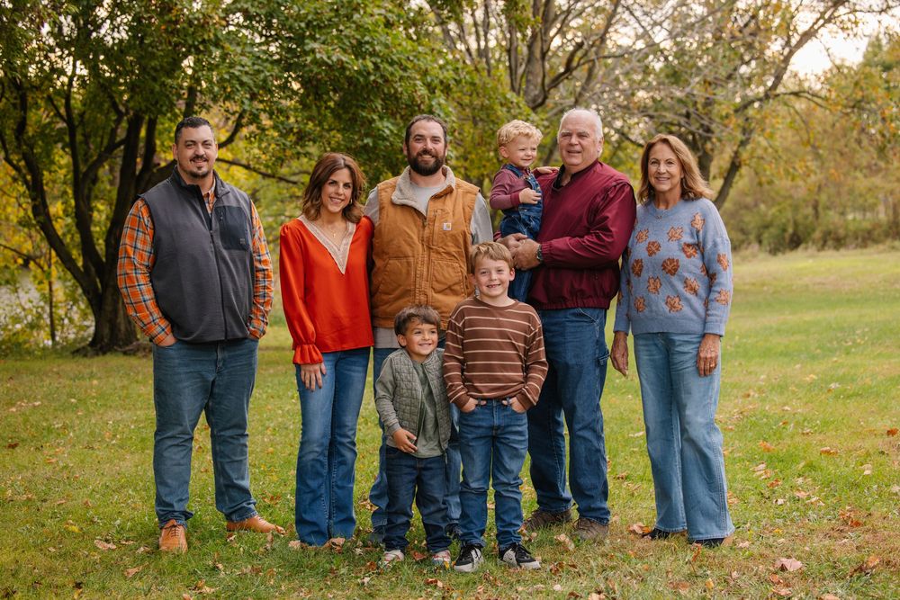 Family of eight posing outdoors in autumn, wearing fall colors, surrounded by trees with colorful foliage.