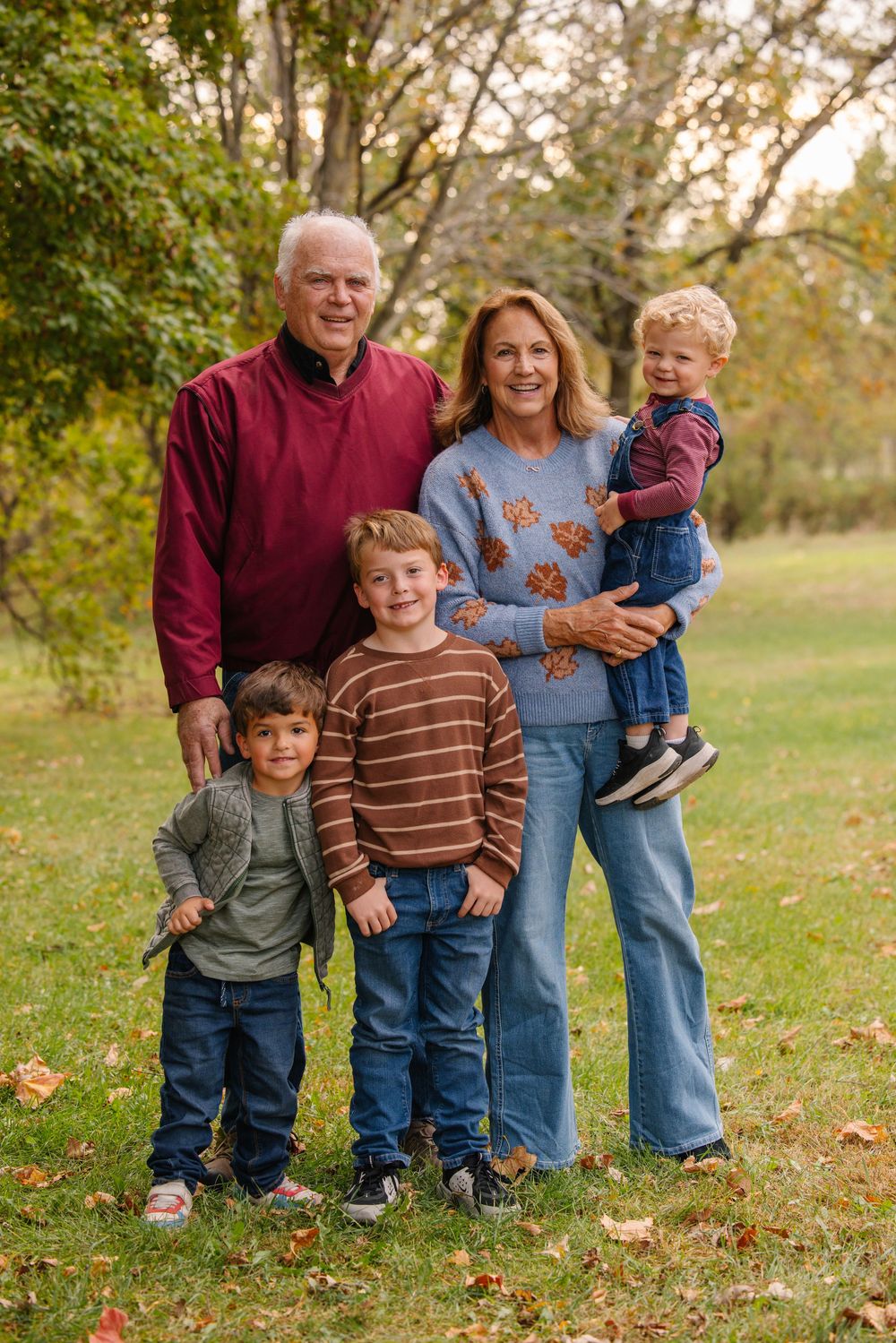 Grandparents pose outdoors in autumn with three young grandchildren on a green lawn with fall foliage.