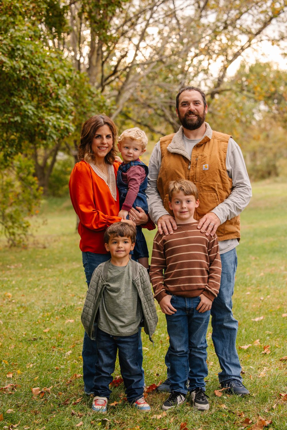 Family of five posing outdoors in fall foliage, wearing autumn colors, smiling for a portrait photo.