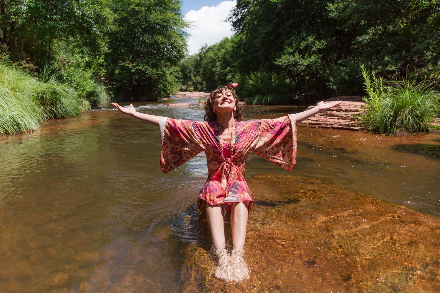 Person in burgundy dress splashing and playing in a shallow creek on a sunny day.