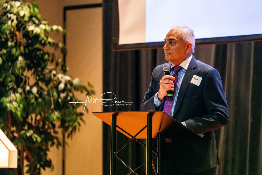 A speaker with white hair and glasses presents at a podium during a professional event.