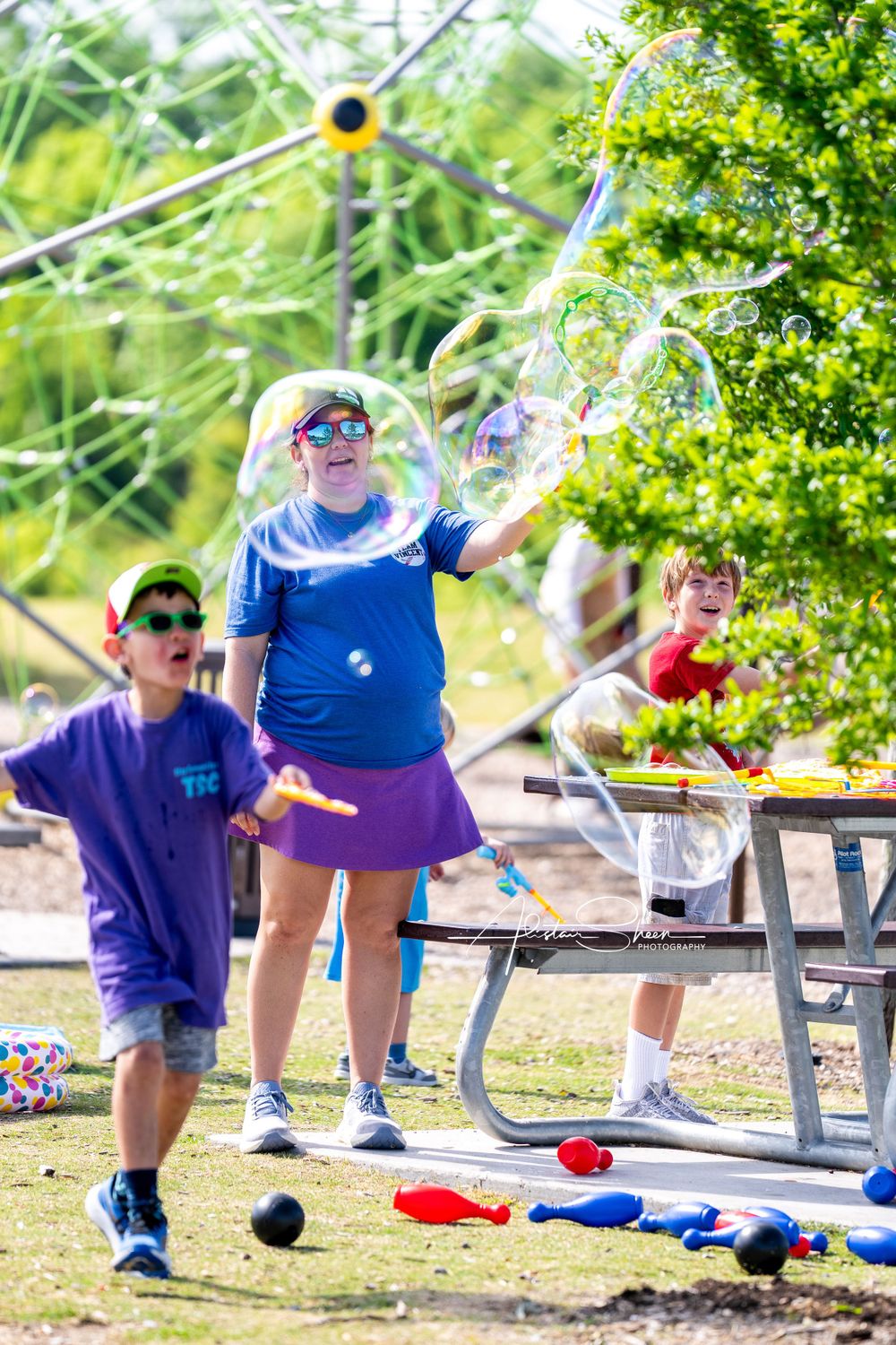 People in colorful clothing play with giant bubbles at an outdoor picnic table on a sunny summer day.