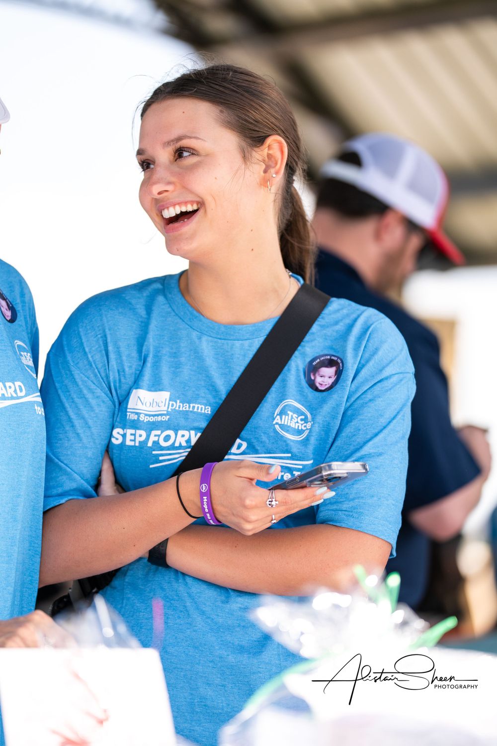 Volunteer in blue t-shirt with event badge smiling while holding phone at outdoor gathering.