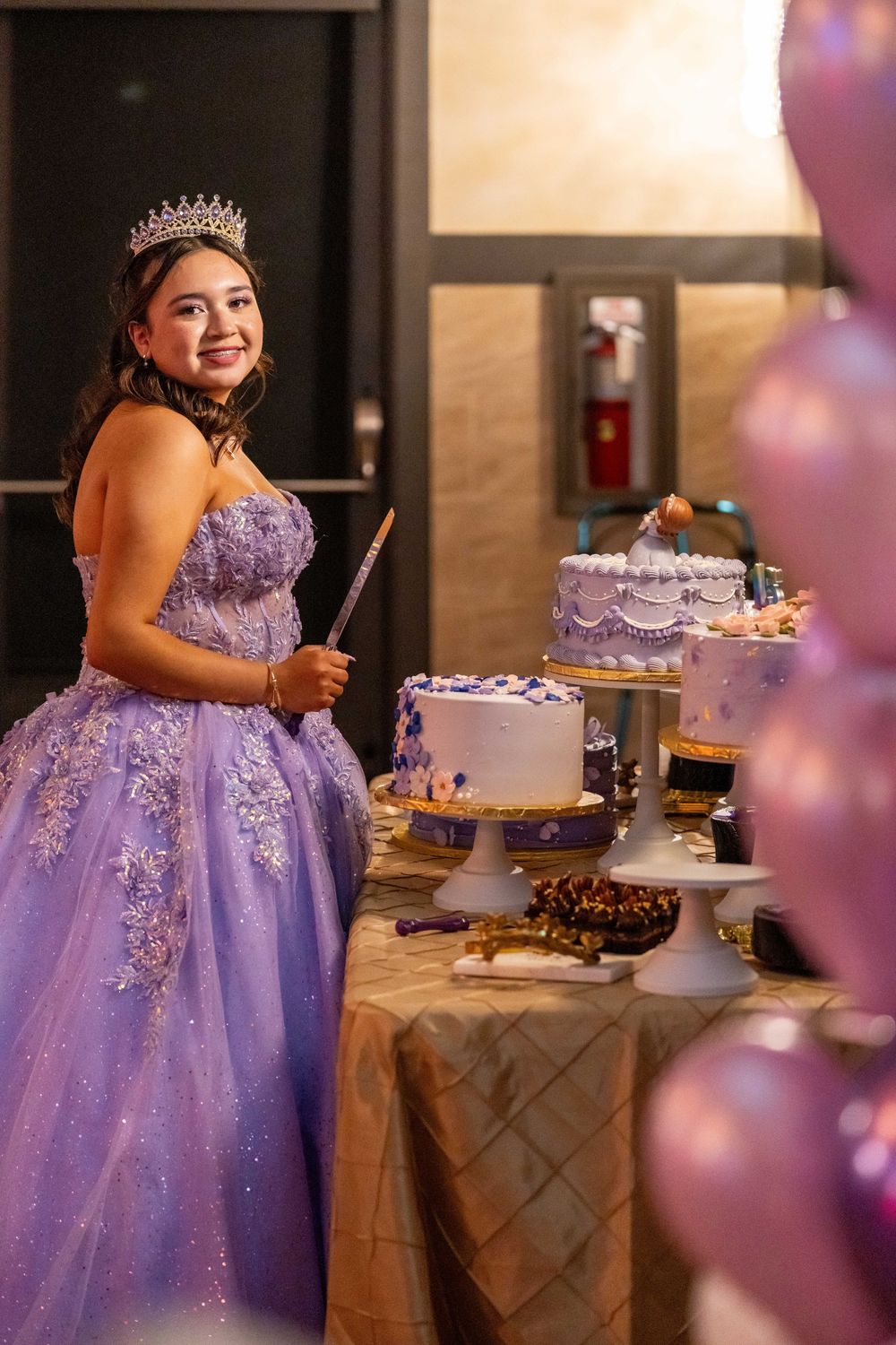 A young person in a lavender quinceañera dress and tiara poses beside an elegant dessert table with cakes.