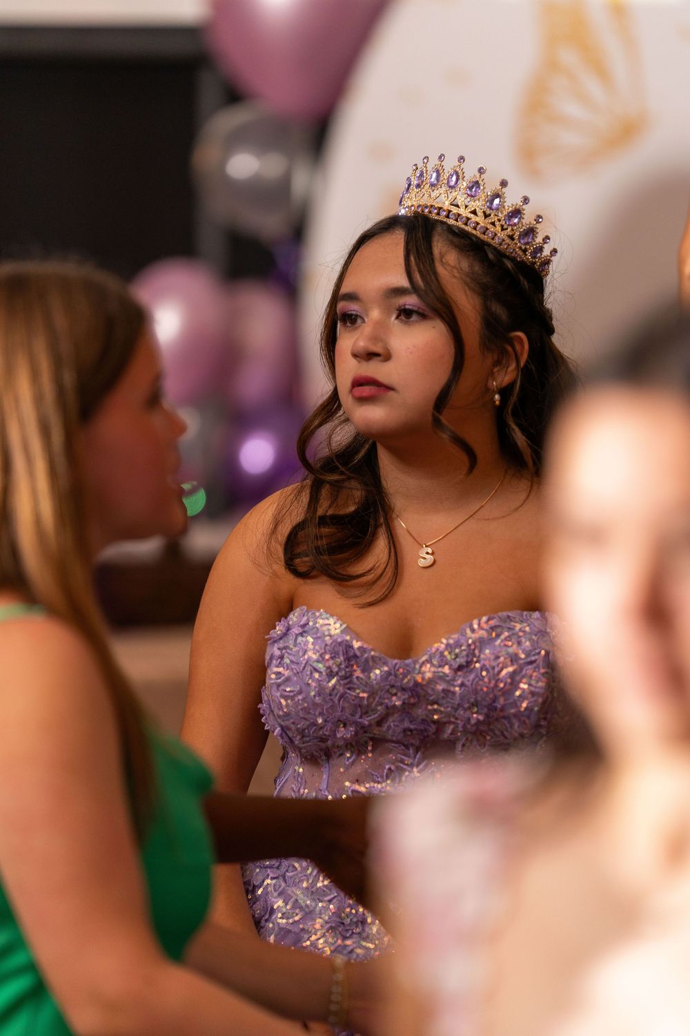A guest in a lavender beaded gown and crystal tiara enjoys a formal celebration with colorful balloons in the background.