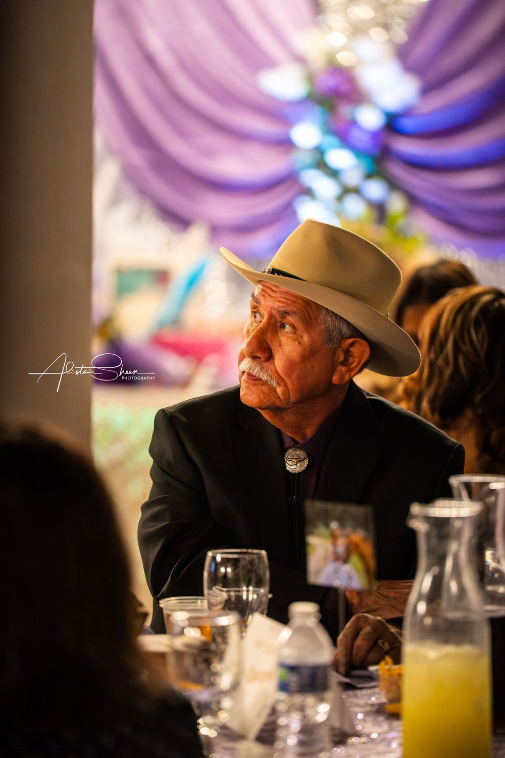 An older man in a cowboy hat sits pensively at a formal dinner table with purple lighting and glassware.