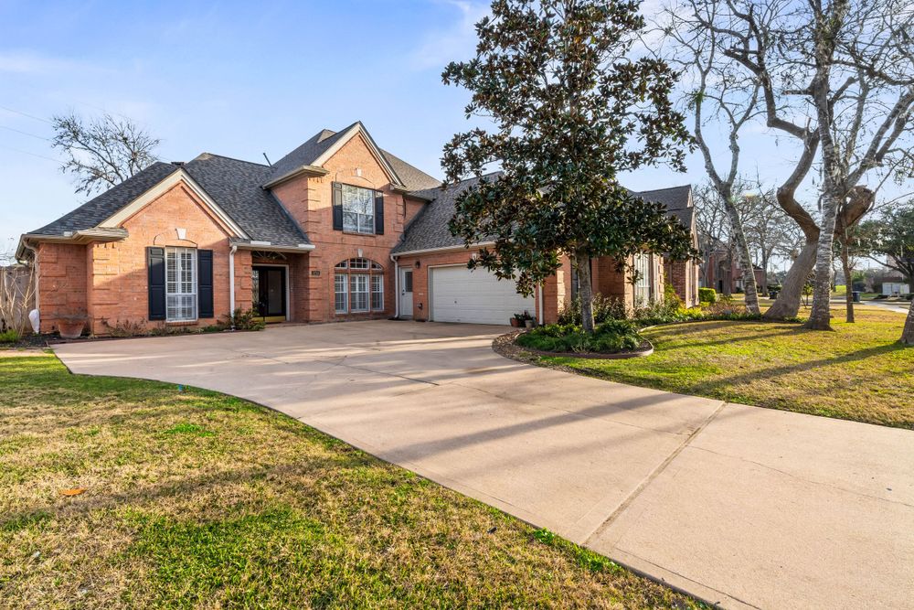 Large brick suburban home with black shutters, arched entryway, and long concrete driveway surrounded by mature trees.