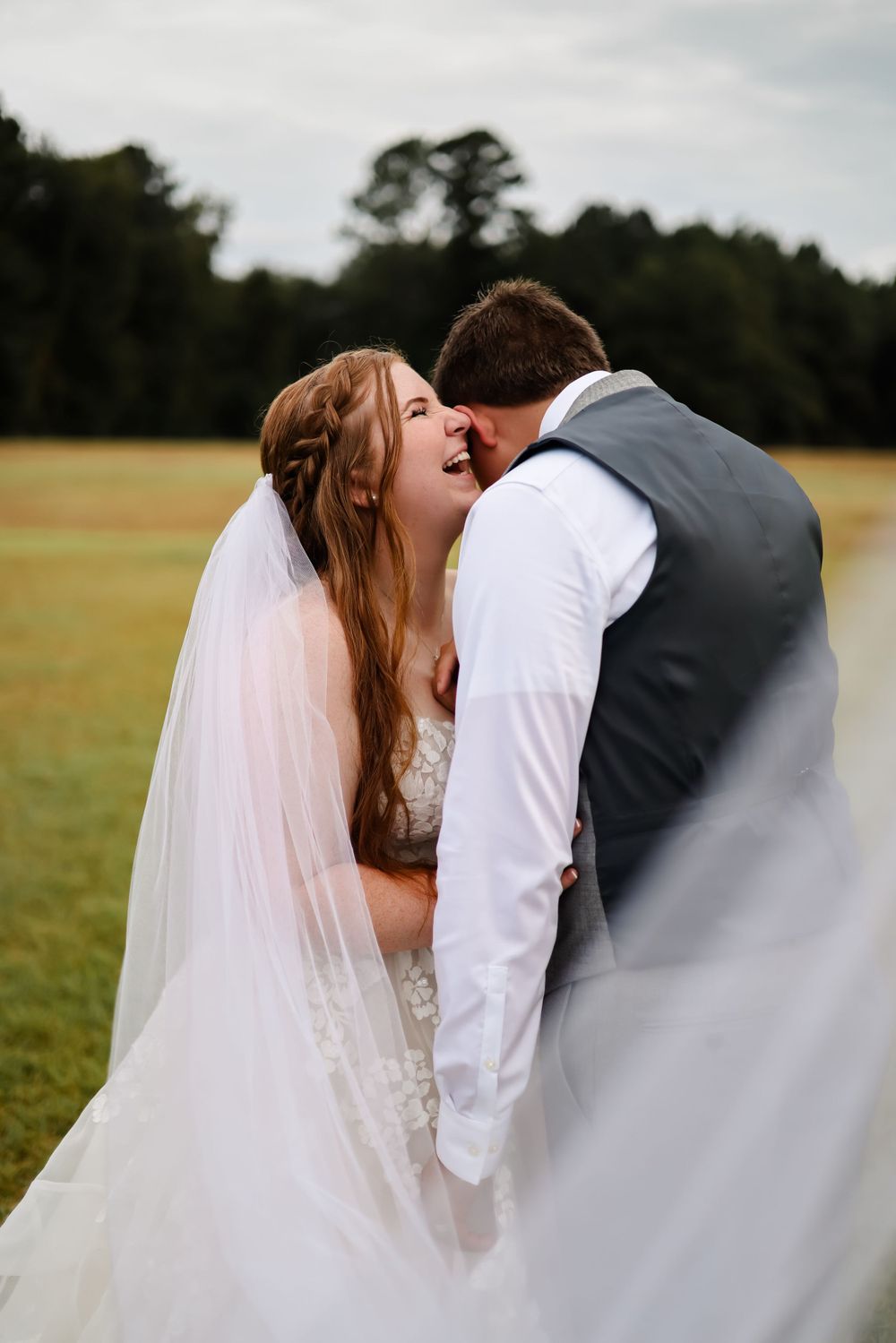 A couple shares a joyful moment on their wedding day in an outdoor field setting with a flowing white veil.