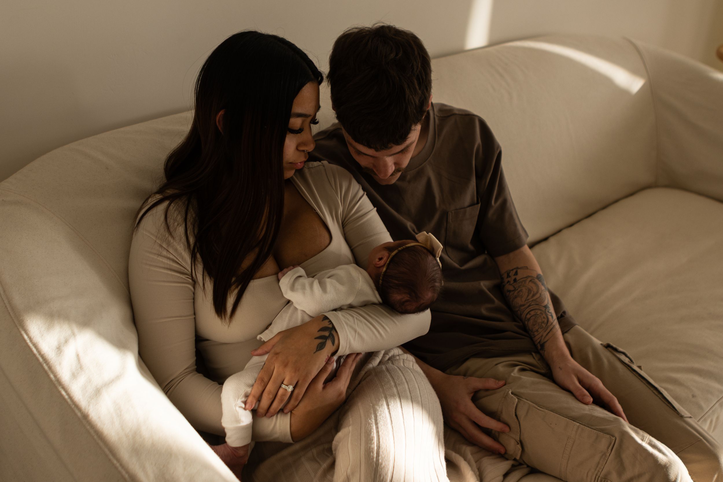 A young couple holds their newborn baby girl on a couch in studio with natural light coming in.