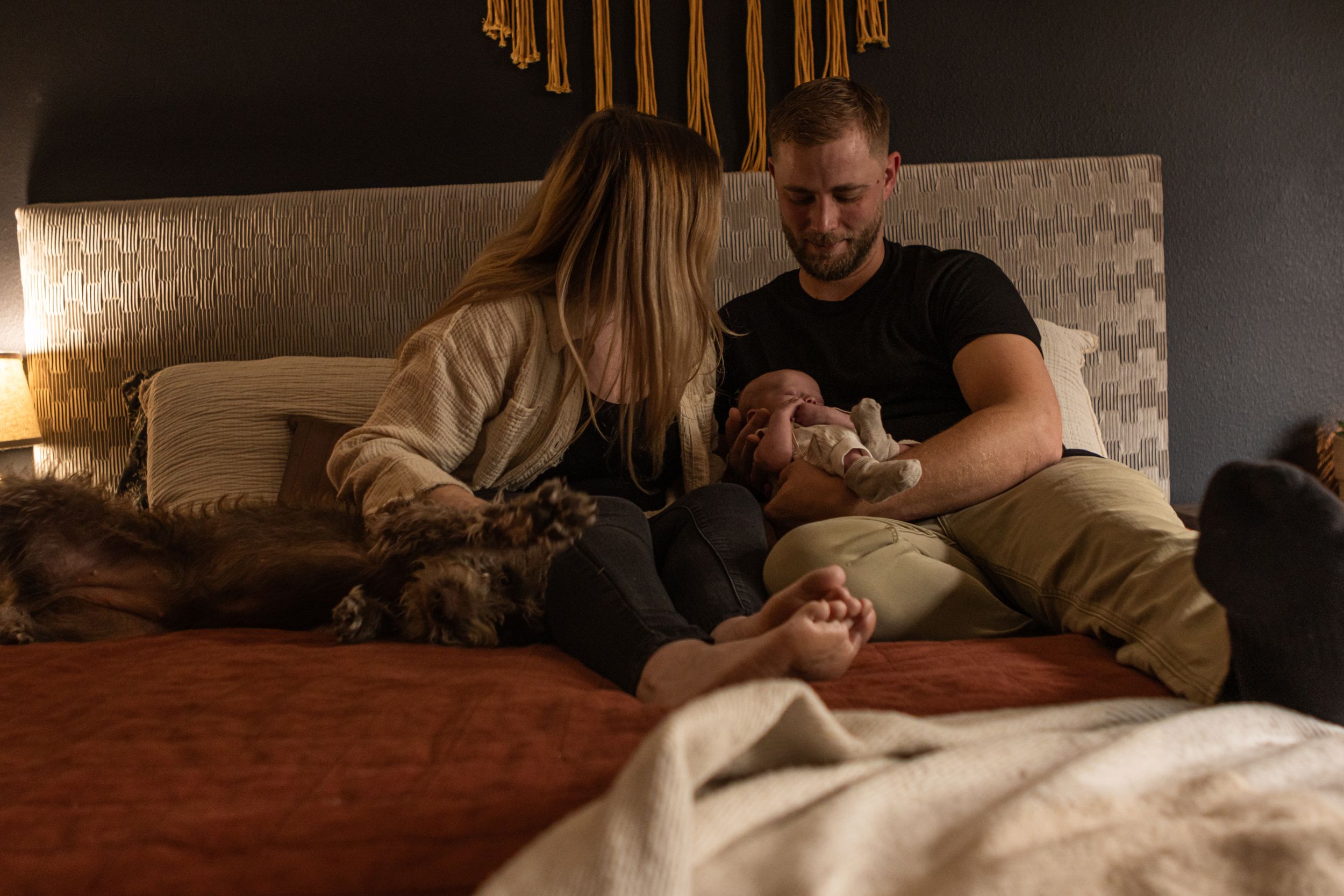 A young family sits on their bed in their bedroom. There is a mom, a dad, a newborn baby boy and their dog