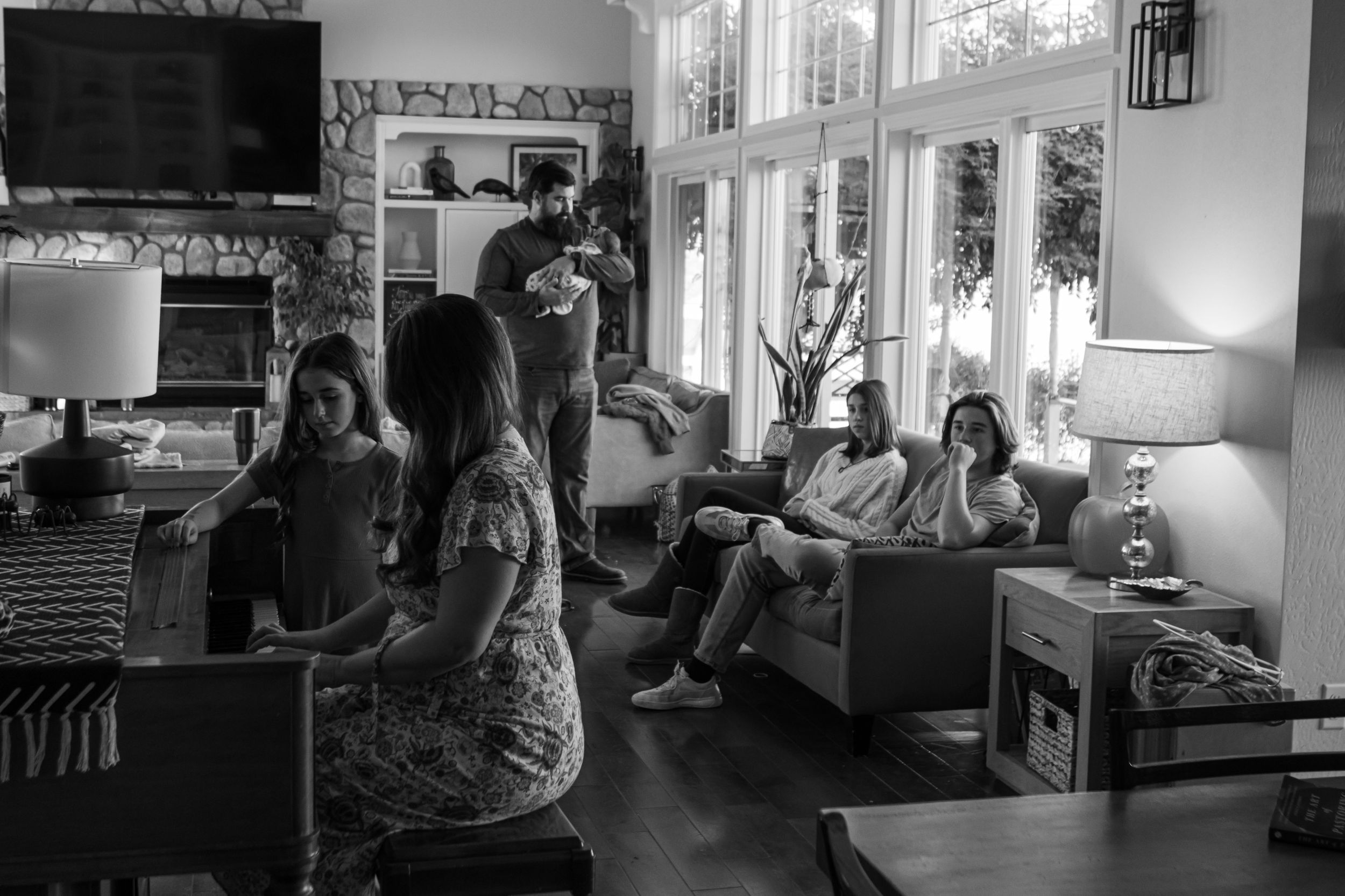 A large family rests in their home photographed in black and white. There is a mom, a dad, two teenagers, a school aged child, and a newborn